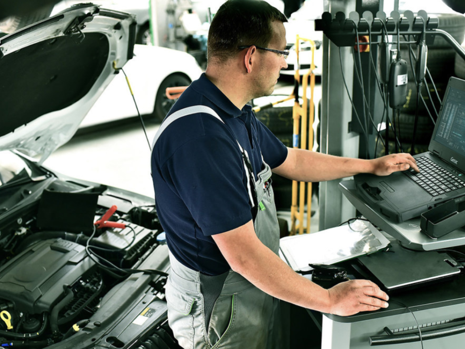 Mecânico automotivo com óculos de proteção realizando diagnóstico eletrônico em um motor com o capô aberto, utilizando um notebook semi-robusto Getac S410 em um carrinho de trabalho em oficina de reparo, o que demonstra o uso de **diagnóstico automotivo robusto** em bancada.